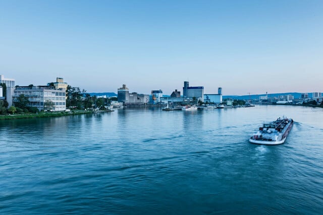 Panorama des Rheinhafens in Basel bei Nacht. Ein Schiff fährt flussaufwärts.