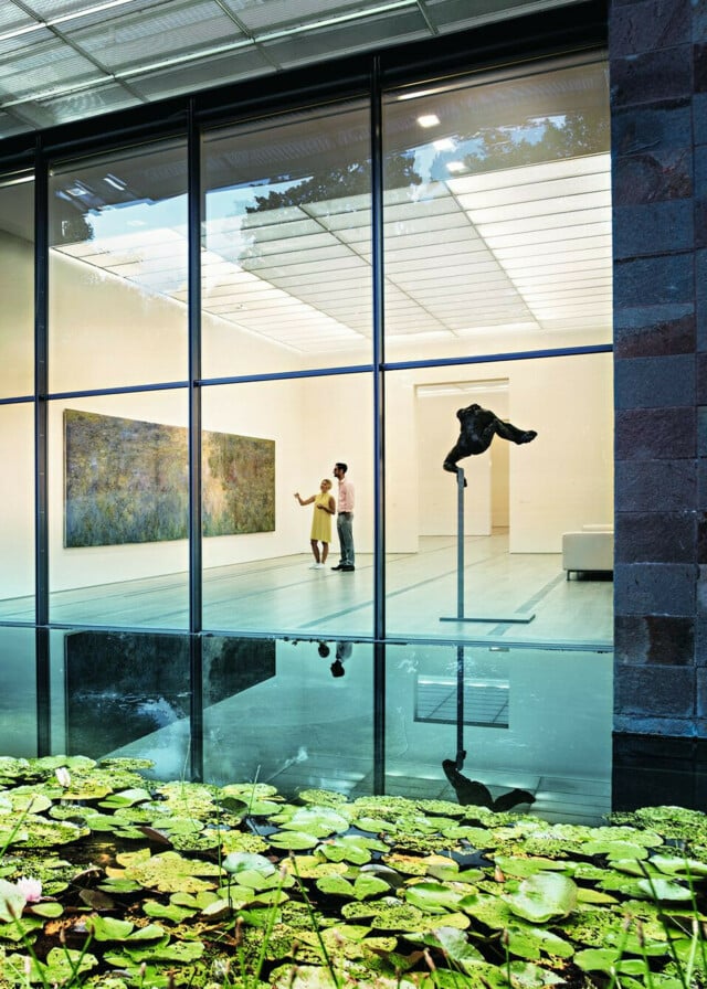 Die Fondation Beyeler in Riehen. Zentral sind Besucher zu sehen, welche das Bild Le Bassin aux nymphéas von Claude Monet betrachten, im Vordergrund der Teich mit Seerosen.