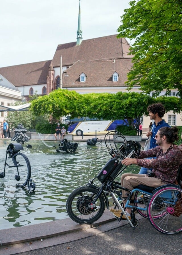 Junger Mann im Rollstuhl mit Begleitung beim Tinguely Brunnen in Basel.